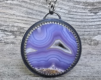 May include: A round pendant necklace featuring a polished purple agate slice with concentric banding and a small central geode cavity. The stone is set in a dark, oxidized metal bezel with a decorative scalloped edge, suspended from a silver chain.