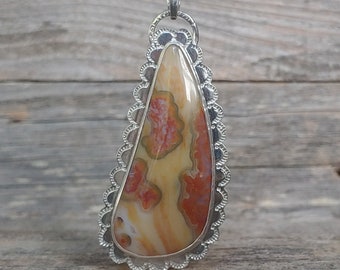 Puede incluir: Un colgante de plata con un borde festoneado, que presenta una piedra en forma de lágrima con un patrón de remolino de naranja, rojo y blanco.