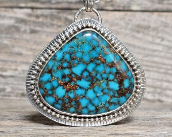 Puede incluir: Un collar con colgante de plata con una gran piedra turquesa triangular. La piedra, con patrones de matriz marrón, está engastada en un bisel de plata detallado con un borde de cuentas. Cuelga de una delicada cadena de plata. El colgante tiene un aspecto artesanal.