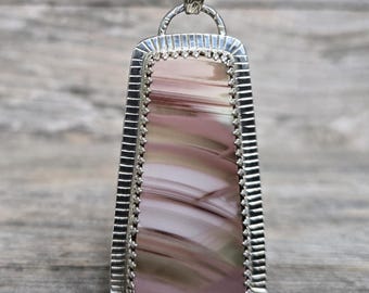 Puede incluir: Un collar con colgante rectangular con una cadena plateada. El colgante presenta una piedra pulida con remolinos rosas, marrones y crema, engastada en un marco plateado con un borde decorativo. El collar se muestra sobre una superficie de madera.