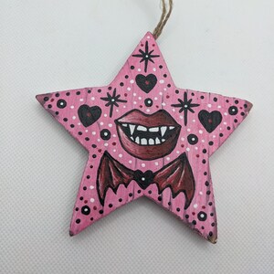 May include: Pink wooden star ornament with a painted vampire face, bat wings, and hearts. The star has a twine hanger.