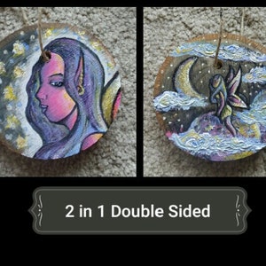 Könnte beinhalten: Doppelseitiger Holzschmuck mit einem gemalten Bild einer Fee mit lila Haaren und einer Sichelmond auf der einen Seite und einer Fee mit Flügeln, die in den Wolken fliegt, auf der anderen Seite. Der Schmuck ist in Schattierungen von Lila, Rosa, Blau und Gold bemalt.