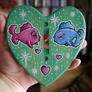 Puede incluir: Una tapa de interruptor de luz en forma de corazón verde con un diseño de peces rosa y azul. Los peces son estilizados y tienen ojos blancos y un contorno blanco. El fondo es verde con puntos blancos y copos de nieve.