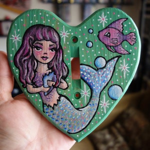 Puede incluir: Una tapa de interruptor de luz en forma de corazón verde con una sirena pintada y un pez rosa. La sirena tiene el pelo morado, una cola azul y sostiene una concha. El fondo es verde con puntos blancos y estrellas.