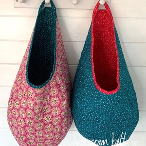 May include: Two hanging fabric storage bins with a teal and pink floral pattern. The bin on the left is pink with a teal trim and the bin on the right is teal with a pink trim. The bins are hanging on white hooks on a white wall. The text "love from beth x" is written on the wall below the bins.