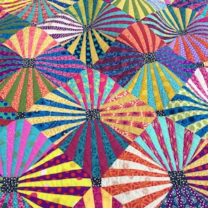 Könnte beinhalten: Ein farbenfroher Quilt aus Stoffquadraten mit einem Sonnenstrahlenmuster. Der Quilt besteht aus verschiedenen Farben, darunter Rot, Gelb, Blau, Grün und Lila.