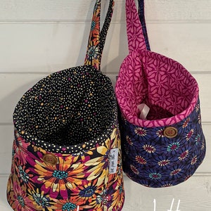 Puede incluir: Dos cestas colgantes de tela con diseños florales. Una presenta un fondo negro con grandes flores amarillas, naranjas y rosas. La otra tiene un fondo azul con flores moradas y amarillas. Ambas tienen botones de madera.