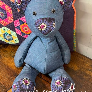 May include: A blue teddy bear with patterned ears, nose, and feet, sitting on a wooden surface. The bear has black button eyes. The text "love from beth x" is written in white cursive. The bear is handmade.