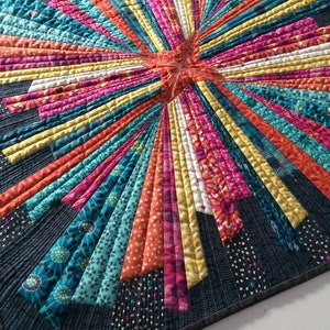 Puede incluir: Una colcha colorida con un patrón de estallido de estrellas en tonos de azul, naranja, amarillo, rosa y blanco. La colcha está hecha de retazos de tela y tiene un borde azul oscuro.