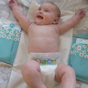 May include: A baby wearing a white diaper with a blue and green cartoon design on the front. The baby is lying on a white changing mat with blue and white patterned changing pads on either side.