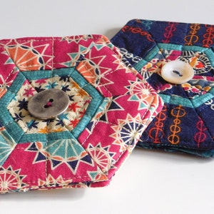 May include: Two handmade, hexagonal coasters. One is magenta with a geometric pattern and a silver button. The other is navy blue with a chain-link design and a white button. Both are quilted and have a teal border.