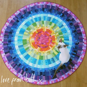 Peut inclure: Un tapis rond coloré avec un motif arc-en-ciel et un chat blanc assis dessus. Le tapis a une bordure violette et un cercle central avec un motif rose et orange. Le texte "love from beth x" est écrit en blanc dans le coin inférieur gauche du tapis.