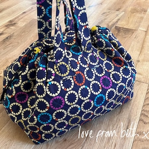 Puede incluir: Bolso tote acolchado azul marino con un patrón repetido de corazones coloridos. El bolso tiene dos asas y un cierre de cordón. El diseño incluye corazones amarillos, naranjas, morados, azules y blancos. El texto "love from beth x" es visible en la parte inferior derecha.