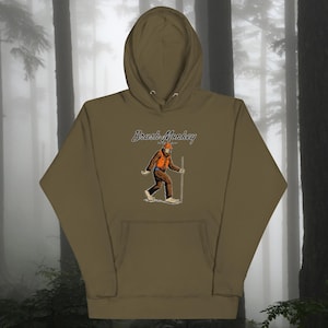 Puede incluir: Una sudadera con capucha verde con el texto "Brush Monkey" y una imagen de dibujos animados de un Bigfoot con un chaleco naranja y un palo.