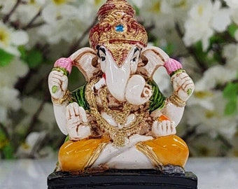 Puede incluir: Una pequeña estatua colorida de la deidad hindú Ganesha. La estatua está hecha de resina pintada y representa a Ganesha sentado con la trompa levantada y sosteniendo un dulce en su mano. La estatua está colocada sobre un fondo de flores blancas.