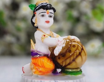 May include: A small, white figurine of a young boy with black hair, wearing a purple and orange outfit, holding a brown pot. The figurine is sitting on a white base with grey stripes.