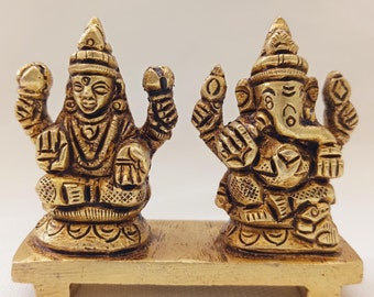 May include: Gold-toned metal statue of a Hindu deity couple, Lakshmi and Ganesha, on a decorative stand. The statues are intricately detailed and have a traditional design.