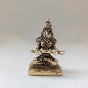 May include: A small, gold-colored brass statue of a seated female figure playing a stringed instrument. The figure is wearing a crown and ornate jewelry. The statue is on a square base.