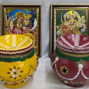 以下が含まれることがあります： 蓋付きの装飾的な壺が2つ、1つは黄色、もう1つは赤で、複雑なデザインと装飾が施されています。壺は、ヒンドゥー教の神々の額縁付きの絵の前に置かれています。
