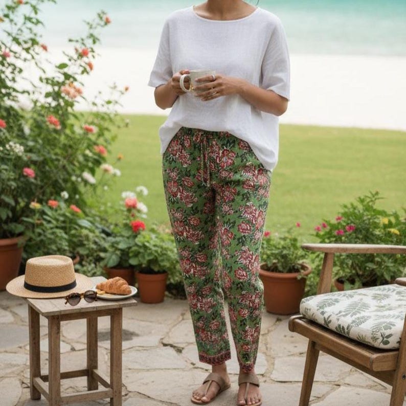 Puede incluir: Una persona con una camisa blanca y pantalones verdes con estampado floral. Un sombrero de paja, gafas de sol y un cruas&aacute;n est&aacute;n sobre una peque&ntilde;a mesa de madera. La persona sostiene una taza. El fondo incluye un jard&iacute;n y vistas al mar.