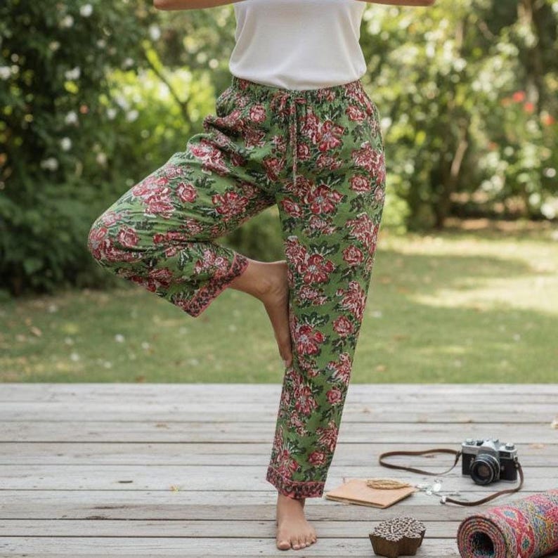 Puede incluir: Pantalones verdes con estampado floral y cintura con cord&oacute;n. Los pantalones presentan un patr&oacute;n repetido de flores rojas sobre un fondo verde. La persona lleva una camiseta blanca y est&aacute; haciendo una postura de yoga en una terraza de madera.