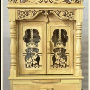 May include: A wooden cabinet with two doors and a drawer. The doors have carved designs of elephants and bells. The cabinet is a light wood color.