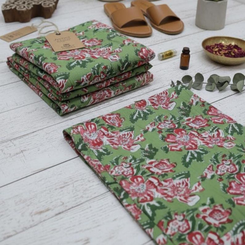 Puede incluir: Servilletas de tela con estampado floral verde con un dise&ntilde;o floral rojo y blanco, apiladas sobre una superficie de madera blanca. Una etiqueta est&aacute; adherida a la servilleta superior. Otros elementos en el fondo incluyen sandalias de cuero marr&oacute;n, aceites esenciales y un cuenco peque&ntilde;o con p&eacute;talos de rosa secos.