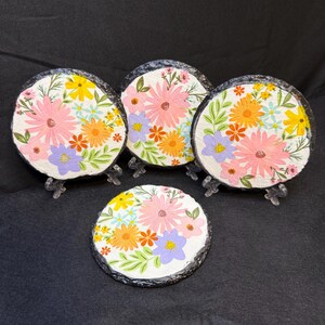 May include: Four round slate coasters with painted floral designs. Each coaster features a different arrangement of pink, orange, yellow, and purple flowers on a white background.
