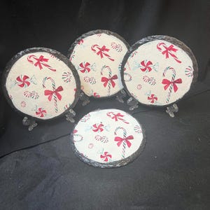 May include: Four round slate coasters with a white background and a repeating pattern of red and white candy canes, red bows, and green and white peppermint candies.