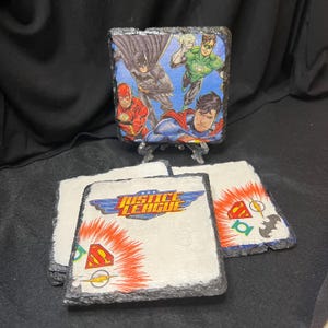 May include: Set of four square coasters featuring Justice League characters. The coasters are made of slate and have a white background with red, blue, and yellow accents. The coasters feature the Justice League logo, Superman, Batman, The Flash, and Green Lantern.