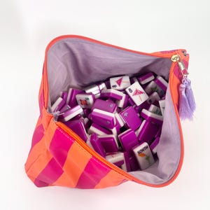 May include: A bright orange and pink striped zippered bag filled with purple and white Mahjong tiles. The bag has a purple interior and a gold zipper with a purple tassel. The tiles have various symbols and numbers.