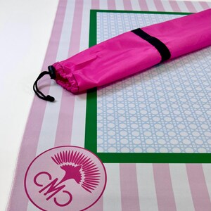 May include: A pink and white striped mat with a green and blue patterned border. A pink carrying bag with a black drawstring is on top of the mat. A pink logo is in the lower left corner.