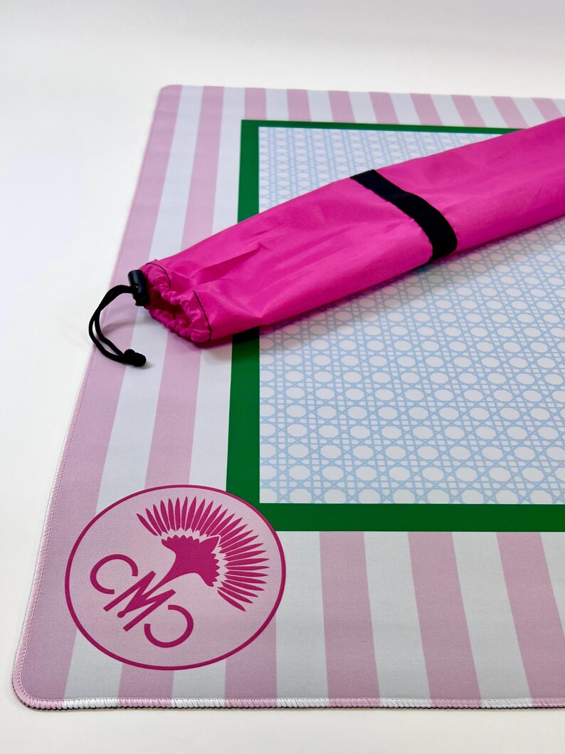 May include: A pink and white striped mat with a green and blue patterned border. A pink carrying bag with a black drawstring is on top of the mat. A pink logo is in the lower left corner.