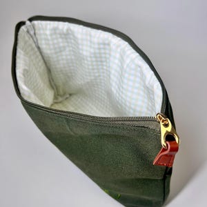 May include: An open, dark green canvas pouch with a gold zipper and a brown leather pull tab. The interior lining features a white and light green gingham pattern. The pouch is designed to hold various items.