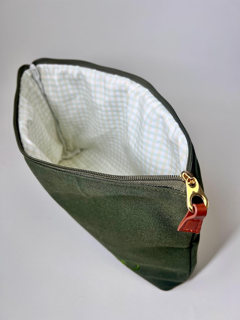 May include: An open, dark green canvas pouch with a gold zipper and a brown leather pull tab. The interior lining features a white and light green gingham pattern. The pouch is designed to hold various items.