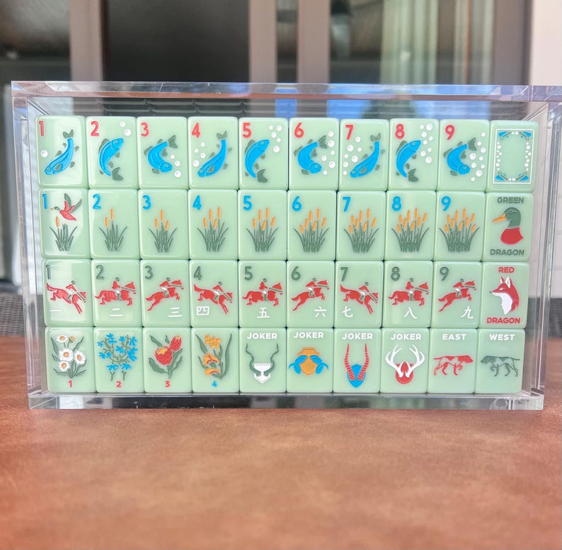 May include: A set of light green Mahjong tiles displayed in a clear acrylic case. The tiles feature various symbols, numbers, and Chinese characters. The set includes tiles labeled "Joker", "East", and "West".