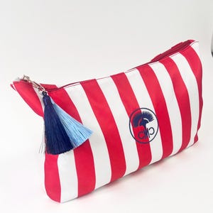May include: Red and white striped cosmetic bag with a navy and light blue tassel. The bag features a navy blue logo on the side. The zipper is silver.