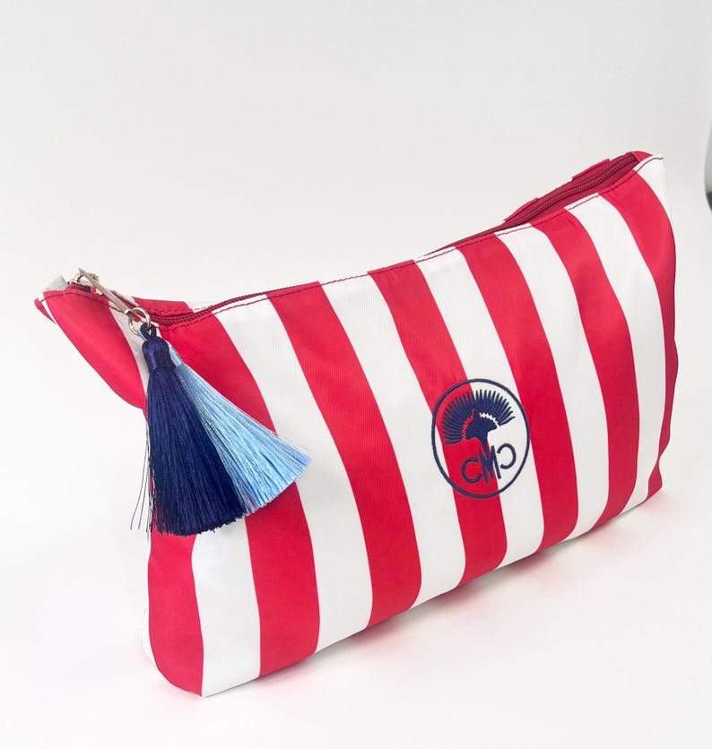 May include: Red and white striped cosmetic bag with a navy and light blue tassel. The bag features a navy blue logo on the side. The zipper is silver.
