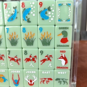 May include: A close-up of a set of green Mahjong tiles with various illustrations and numbers. The tiles feature images of fish, reeds, ducks, foxes, and horse riders. Some tiles have the words "JOKER", "EAST", "WEST", "GREEN", and "RED DRAGON".