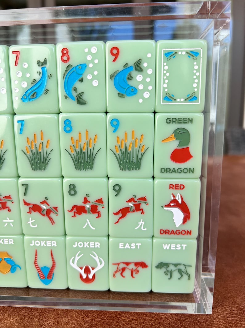 May include: A close-up of a set of green Mahjong tiles with various illustrations and numbers. The tiles feature images of fish, reeds, ducks, foxes, and horse riders. Some tiles have the words "JOKER", "EAST", "WEST", "GREEN", and "RED DRAGON".