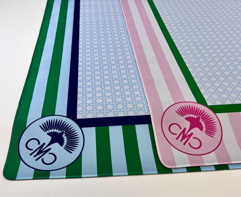 May include: Two decorative mats with striped and patterned designs. One mat features green and light blue stripes with a blue and white logo. The other mat has pink and white stripes with a pink and white logo. Both mats have a light blue patterned center.