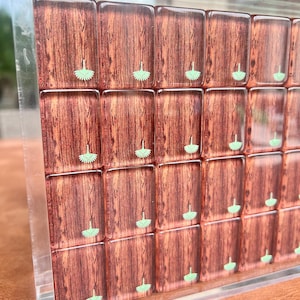 May include: A close-up of a decorative wooden game board with a clear acrylic frame. The board features rectangular tiles with a wood grain pattern and small green floral designs. The board is displayed at an angle.