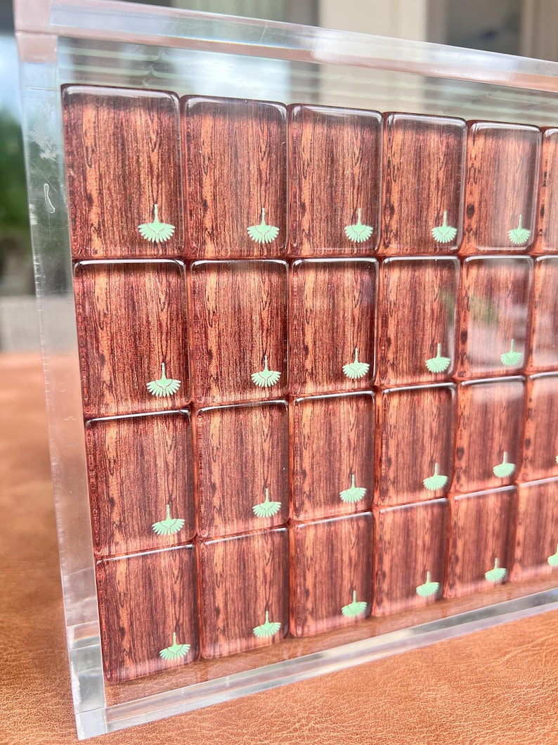 May include: A close-up of a decorative wooden game board with a clear acrylic frame. The board features rectangular tiles with a wood grain pattern and small green floral designs. The board is displayed at an angle.