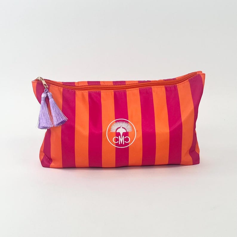 May include: A rectangular cosmetic bag with vertical stripes in bright pink and orange. The bag has a gold zipper and two lavender tassels. A white circular logo with the letters "CMO" is centered on the bag.