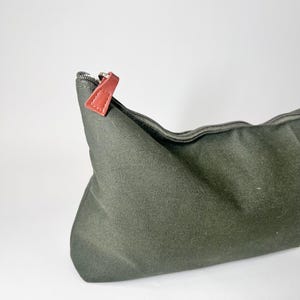 May include: An olive green canvas cosmetic bag with a zipper closure. The zipper pull is a reddish-brown leather. The bag is empty and has a simple, minimalist design. The bag is on a white background.
