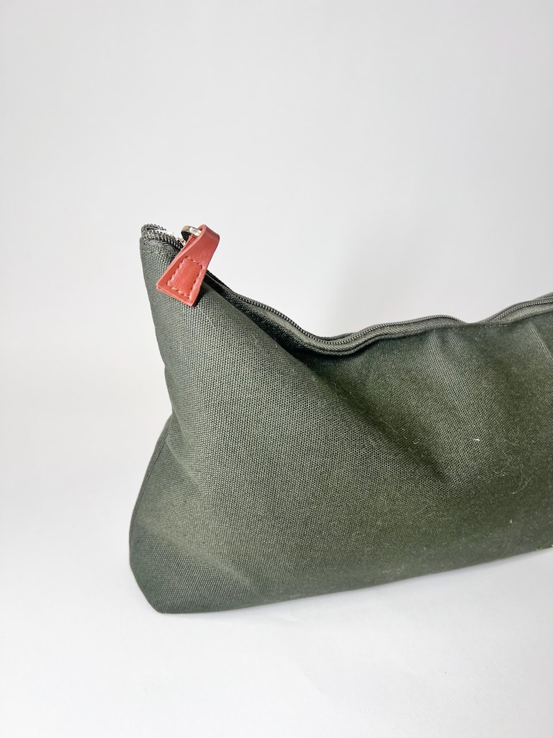 May include: An olive green canvas cosmetic bag with a zipper closure. The zipper pull is a reddish-brown leather. The bag is empty and has a simple, minimalist design. The bag is on a white background.