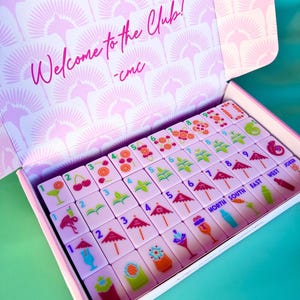 May include: A pink and white Mahjong set in a box. The box lid reads "Welcome to the Club!" in pink script. The tiles are light pink with colorful illustrations of cherries, umbrellas, and cocktails. The set includes tiles with numbers and directional markers.