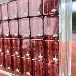 May include: A clear acrylic box filled with rectangular wooden blocks. Each block has a rich, reddish-brown wood grain and a small, light green fan-shaped design. The blocks are arranged in a grid pattern, creating a visually appealing display.