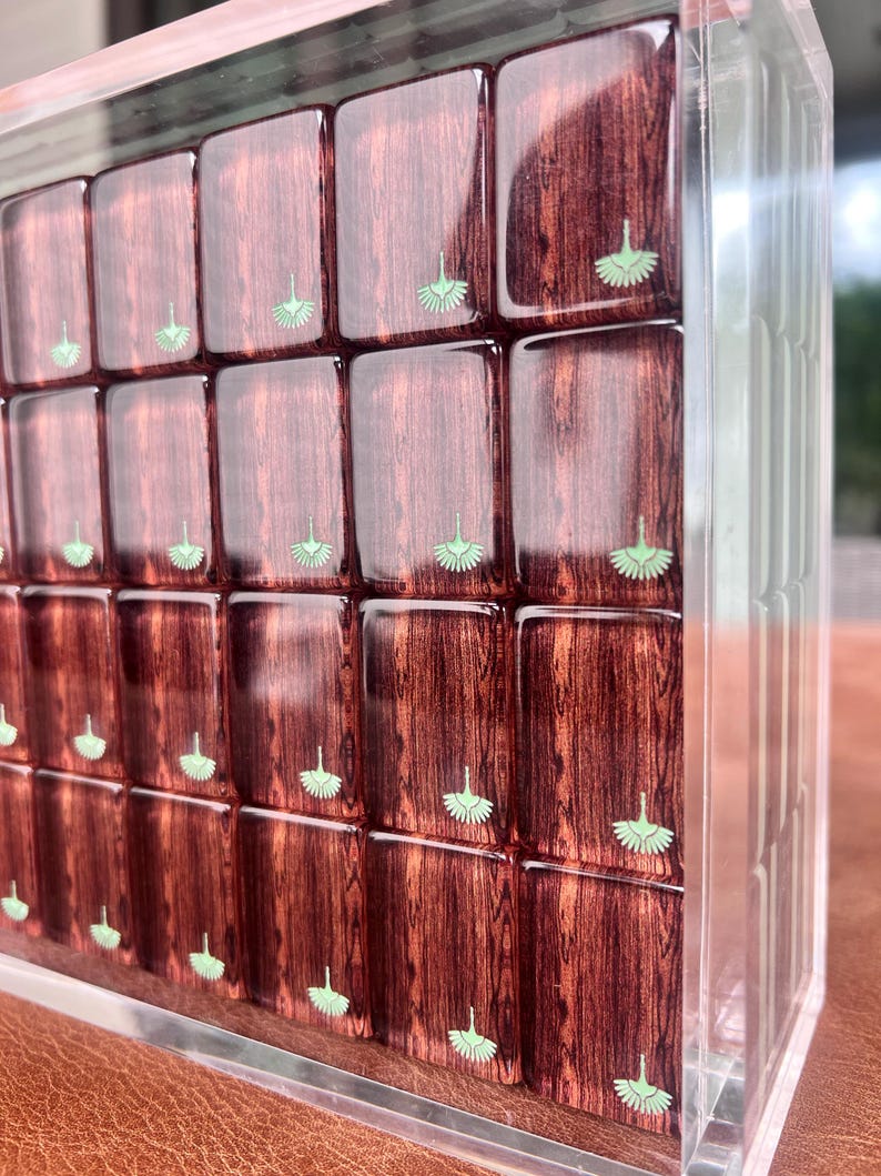 May include: A clear acrylic box filled with rectangular wooden blocks. Each block has a rich, reddish-brown wood grain and a small, light green fan-shaped design. The blocks are arranged in a grid pattern, creating a visually appealing display.