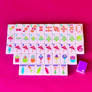 May include: A set of white Mahjong tiles with colorful illustrations of cocktails, fruit, and plants. The tiles are arranged on a bright pink surface, with a small purple tile box. The text "NORTH SOUTH EAST WEST JOKER JOKER" is visible.